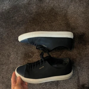 Axel Arigato Cap Toe Sneakers - Snygga svarta sneakers från Axel Arigato med vit sula och diskret guldtext på sidan. Skorna har snörning och en stilren design som passar perfekt till vardags. Liknar lanvin cap toe, mörkblå, ej svarta. En väldigt liten fläck också som syns på sista bilden, går nog bort om man tvättar.