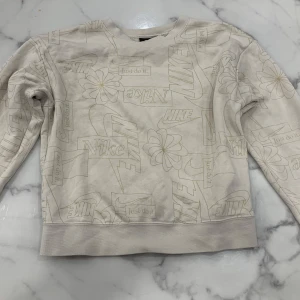 Beige sweatshirt från Nike - Säljer en beige sweatshirt från Nike med ett coolt mönster av blommor och logotyper. Tröjan har långa ärmar och en avslappnad passform. 