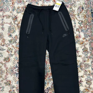 Nike Tech Fleece (Helt ny)  - Säljer ett par utsläppta svarta mjukisbyxor från Nike med dragsko i midjan och praktiska dragkedjefickor. Perfekta för en avslappnad stil eller träning. Byxorna har en diskret Nike-logga på benet.