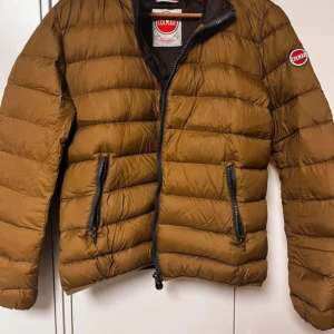 Brun dunjacka från Colmar - Snygg brun dunjacka från Colmar med dragkedja och två praktiska fickor framtill. Jackan har en quiltad design och är perfekt för kyligare dagar. Jackan är i nyskick och kostar ca 3000 som ny