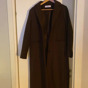 Brun kappa från Ellos - Snygg brun kappa från Ellos i storlek 38. Den har en klassisk design med långa ärmar och knappar framtill. Perfekt för höst och vinter, tillverkad i en blandning av polyester och ull för en bekväm passform.