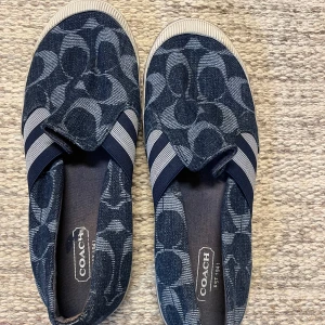  Coach slipon sko i blå denimcanvas, stl 36,5 - Jeansblå med coachsignaturen och läderdetalj på baksidan, elastiskt band, lite töjbara framtill, gummisulor. Stl 36,5, normala i storleken.  Använda några ggr, i fint skick, finns lite märken på det vita gummit på sidorna (ej provat ta bort) 
