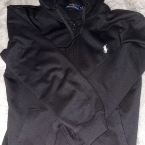 Svart hoodie från Ralph Lauren - Säljer en stilren svart hoodie från Ralph Lauren. Den har en klassisk design med dragkedja och den ikoniska loggan på bröstet. Perfekt för både vardag och chill. Passar till allt och är superbekväm! Stl M men passar S! Kom dm vid frågor eller funderingar!😊