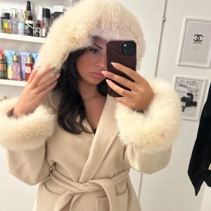 Beige pälsjacka med huva - Säljer en supermysig beige pälsjacka med fluffig huva och ärmslut. Jackan har ett knytbälte i midjan för en snygg passform. Perfekt för kyliga dagar när du vill känna dig både varm och stilren. Passar perfekt till höst och vinter!