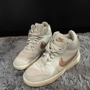 Nike sneakers i beige och brunt - Säljer ett par snygga Nike sneakers i beige med bruna detaljer. Skorna har en hög modell med snörning och är i bra skick. Perfekta för både vardag och träning. Storlek dam 39.