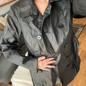 Svart trenchcoat från Vero Moda - Säljer en snygg svart trenchcoat från Vero Moda i storlek XL. Jackan har en klassisk design med knappar framtill och långa ärmar. Perfekt för höst och vår, och ger en stilren look till vilken outfit som helst. Passar både till vardags och lite finare tillfällen.