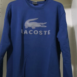 Blå tröja från Lacoste - Säljer en snygg blå tröja från Lacoste med deras ikoniska krokodillogga på bröstet. Tröjan är långärmad och perfekt för både vardag och chill. Den har en normal passform och är superbekväm. Perfekt för höst och vår!