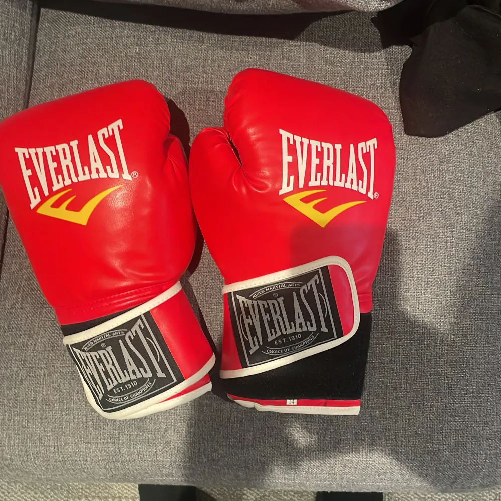 Säljer ett par röda boxningshandskar från Everlast. De är i skinn och har en klassisk design med vit Everlast-logga och gula detaljer. Handskarna har en bred kardborrestängning för extra stöd och ventilation på insidan för komfort. Perfekta för träning eller sparring! 🥊. Övrigt.