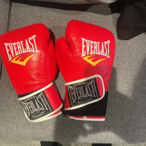 Röda boxningshandskar från Everlast - Säljer ett par röda boxningshandskar från Everlast. De är i skinn och har en klassisk design med vit Everlast-logga och gula detaljer. Handskarna har en bred kardborrestängning för extra stöd och ventilation på insidan för komfort. Perfekta för träning eller sparring! 🥊