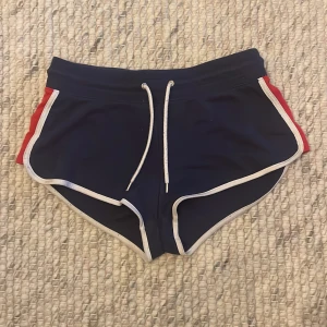Mörkblå shorts med vita och röda detaljer - Säljer ett par mörkblå shorts med vita kanter och röda sidopaneler. De har en bekväm passform med snörning i midjan för justering. Perfekta för sommarens alla aktiviteter!