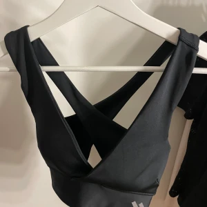Svart sporttopp med korsade band - Säljer en snygg svart sporttopp med korsade band i ryggen. Toppen är perfekt för träning och har en stilren design med en liten vit logga på framsidan. Materialet känns mjukt och stretchigt, vilket ger en bekväm passform.