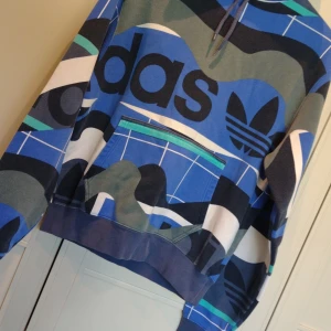 Flerfärgad hoodie från Adidas - Säljer en supercool flerfärgad hoodie från Adidas. Den har ett unikt mönster med blå, grön och vit färg och stora Adidas-loggor. Perfekt för att sticka ut i mängden! Den har en stor ficka fram och en bekväm huva med snörning.