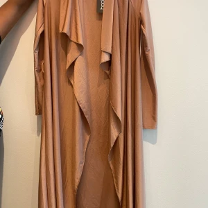 Beige långklänning från Boohoo - Säljer en superfin beige lång kimono från Boohoo. Den har en elegant och flytande design med långa ärmar och en öppen front som ger en snygg look. Perfekt för både vardag och festliga tillfällen!