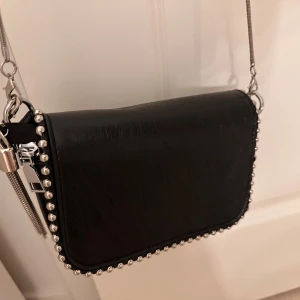 Svart axelväska med nitar - Säljer en supercool svart axelväska med nitar längs kanterna. Den har en stilren design med en silverfärgad kedja som axelrem. Perfekt för en utekväll eller när du vill lägga till lite edge till din outfit!
