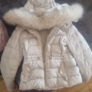 Vit dunjacka från Zara - Säljer en superfin vit dunjacka från Zara Woman. Den har en mysig fuskpäls på huvan och är perfekt för kalla vinterdagar. Jackan har en dragkedja framtill och praktiska fickor med knappar. Insidan är fodrad med mjukt material för extra värme. Perfekt för både vardag och festliga tillfällen!