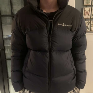 Sail racing Down jacket - Jätte varm och skön sail racing jacka. Inbyggd vind luva i kragen, vatten avvisande och vind tät Den har 2 fickor framtill och en på insidan. Mössa medföljer. Som ny och har inga skador. Ändast använd förra säsongen. Säljer på grund av att ska köpa n