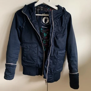 Mörkblå jacka från Elvine - Säljer en snygg mörkblå jacka från Elvine i storlek XS. Jackan har en dragkedja framtill och ribbade muddar vid ärmslut och nederkant. Den har en huva och praktiska fickor med lock.  Perfekt för höst och tidig vinter! 