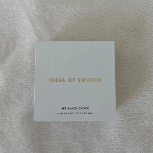 Jet Black Croco AirPods-väska från iDeal of Sweden - Säljer en superstylish Jet Black Croco AirPods-fodral från iDeal of Sweden. Den är i svart med ett croco-mönster och har en snygg guldfärgad kedja. Perfekt för att hålla dina AirPods säkra och trendiga! Passar både 1:a och 2:a generationens AirPods. 