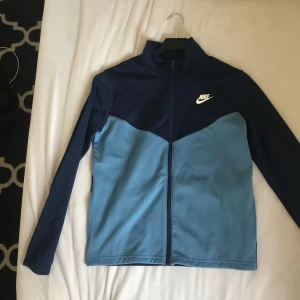 Blå träningsjacka från Nike - Säljer en snygg blå träningsjacka från Nike. Den har en dragkedja framtill och är långärmad. Perfekt för träning eller en avslappnad stil. Jackan är i bra skick och har en stilren design med Nike-loggan på bröstet.
