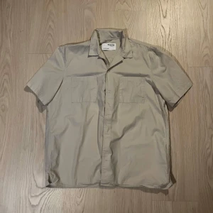 Kortärmad Skjorta - Snygg beige skjorta från Maison Kitsuné med korta ärmar och klassisk krage. Skjortan har två bröstfickor och en stilren design.