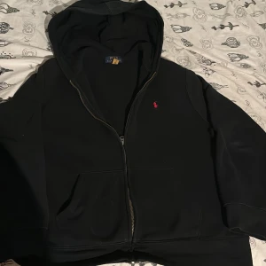 Svart hoodie från Ralph Lauren - Säljer en klassisk svart hoodie från Ralph Lauren med dragkedja och broderad röd logga på bröstet. Perfekt för en avslappnad stil. Hoodien har en bekväm passform och är tillverkad i mjukt material. Pris kan diskuteras 