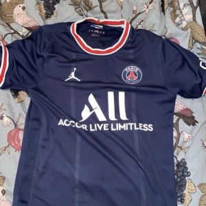 Mörkblå PSG fotbollströja - Snygg mörkblå PSG fotbollströja med korta ärmar och vita samt röda detaljer vid krage och ärmslut. Tröjan har PSG-loggan och Jordan-märket på bröstet samt texten 'Accor Live Limitless' på framsidan.