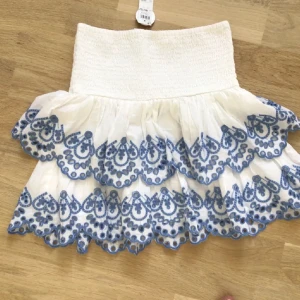 Vit kjol med blå broderier - Charmig vit kjol med vackra blå broderier och volanger. Den har en elastisk midja för bekväm passform och är perfekt för en somrig look. Kjolen är kort och har en luftig design.