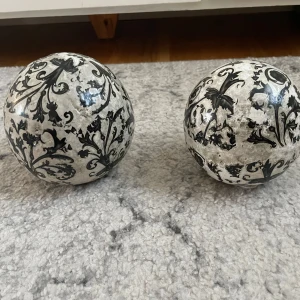 Dekorativa klot med svart mönster - Två dekorativa klot med ett intrikat svart mönster på en vit bakgrund. Kloten har en antik look och är perfekta som inredningsdetaljer. De har små filtkuddar på undersidan för att skydda ytor.