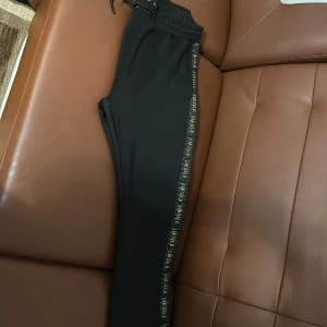 Svarta mjukisbyxor från SikSilk - Snygga svarta mjukisbyxor från SikSilk med elastisk midja och snörning. Byxorna har en diskret logga längs sidan och är perfekta för en avslappnad stil. De är tillverkade i ett mjukt material som ger en bekväm passform.