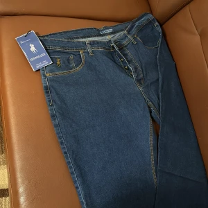 Mörkblå jeans från New P010 ASSN - Snygga mörkblå jeans med klassisk design från New P010 ASSN. De har en rak passform och är tillverkade i slitstarkt denim. Byxorna har knappar och dragkedja framtill samt fem fickor. Perfekta för en stilren look.