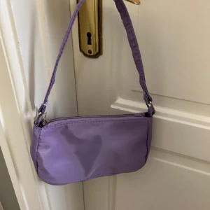 Lila axelväska - Snygg lila axelväska i syntetmaterial med praktisk dragkedja. Perfekt för att bära med sig det viktigaste på språng. Enkel och stilren design som passar till många tillfällen.