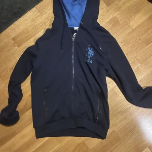 Mörkblå hoodie från U.S. Polo Assn. - Säljer en mörkblå hoodie från U.S. Polo Assn. med dragkedja och broderad logga på bröstet. Insidan av huvan är ljusblå och jackan har långa ärmar. Perfekt för en avslappnad stil.