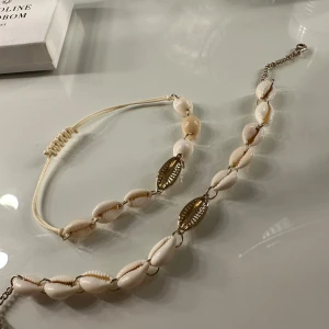 Vitt snäckskalshalsband och armband - Säljer ett set med ett halsband och ett armband dekorerade med vita snäckskal och guldfärgade detaljer. Perfekt för en somrig look! Halsbandet har en justerbar kedja och armbandet har en elastisk tråd för enkel passform.