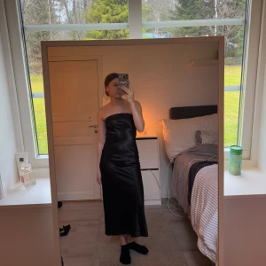 Svart axelbandslös klänning - Snygg svart axelbandslös klänning i satin med en elegant öppen rygg. Perfekt för en kväll ute eller ett speciellt tillfälle. Klänningen har en tajtare passform och når ner till vaderna.