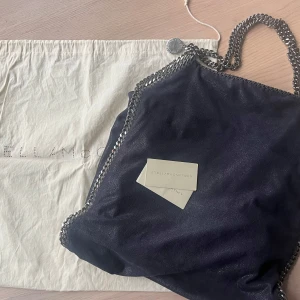 Mörkblå axelväska från Stella McCartney - Snygg svart axelväska från Stella McCartney med kedjedetaljer och dragkedja. Väskan har en insida i en kontrasterande färg och är perfekt för att bära dina dagliga nödvändigheter. Den har en stilren design som passar till många olika outfits. Den är knappt använd, vintage. Stor tote.