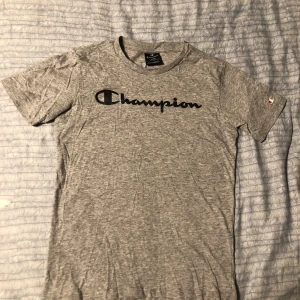 Grå t-shirt från Champion - Säljer en grå t-shirt från Champion med deras ikoniska logga tryckt i svart på bröstet. T-shirten är kortärmad och tillverkad i mjukt bomullsmaterial. Perfekt för en avslappnad stil.