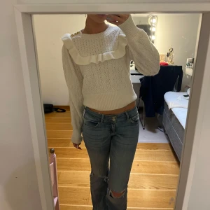 Vit stickad tröja med volangdetalj - Säljer en söt vit stickad tröja med volangdetalj runt halsen. Tröjan har långa ärmar och en ribbad struktur som ger en fin passform. Perfekt för kyligare dagar!