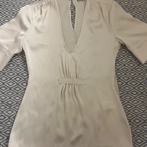 Beige blus från Reiss - En väldigt fin  beige blus från Reiss med v-ringning och korta ärmar. Den har en dragkedja vid sidan och en knapp vid nacken. Den har andvänts fåtals gånger och är i ett väldigt bra skick!