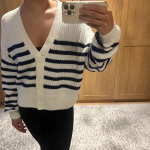 Vit och blå randig kofta - Säljer en snygg vit kofta med blå ränder. Den har en djup v-ringning och stora knappar framtill. Perfekt för en avslappnad stil.