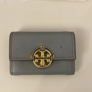 Grå plånbok med gulddetaljer - Tory Burch plånbok. Plånboken har en praktisk dragkedja för myntfacket och en knappstängning. Perfekt för att hålla ordning på kort och kontanter. Den är köpt för 1400kr, priset kan diskuteras 
