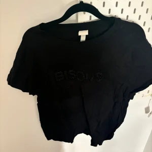Svart  t-shirt med text - Snygg svart croppad t-shirt med texten 'BISOUS' i glittriga detaljer. Perfekt för en avslappnad stil. Kortärmad och tillverkad i mjukt material för extra komfort.