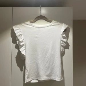 Vit ribbad topp från Shein - Säljer en söt vit ribbad topp från Shein med volangdetaljer vid ärmarna. Perfekt för en stilren och trendig look. Toppen har en enkel design som gör den lätt att matcha med olika outfits.