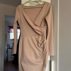 Beige omlottklänning från NLY One - Snygg beige omlottklänning från NLY One i storlek S. Klänningen har en v-ringad design och långa ärmar, perfekt för en stilren look. Den har en draperad detalj i midjan som ger en smickrande passform.