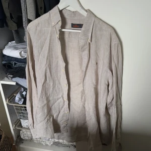 Beige linneskjorta från Morris - Säljer en stilren beige linneskjorta från Morris. Skjortan har långa ärmar och knappar framtill. Perfekt för en avslappnad men ändå elegant look. Den har en liten broderad detalj på bröstet som ger extra stilpoäng.