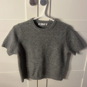 Grå stickad tröja från Zara - Säljer en stilren grå stickad tröja från Zara.Tröjan har korta ärmar och en rund halsringning, perfekt för lager-på-lager under kyligare dagar. Den är mjuk och bekväm, vilket gör den till ett mångsidigt tillskott i garderoben. Passar bra till både jeans och kjol.