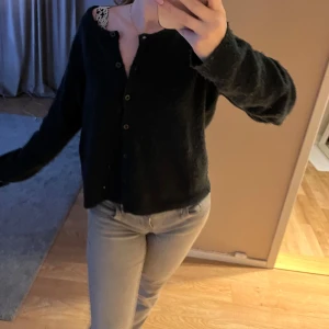 Svart kofta med spetsdetalj - Mysig svart kofta med knappar framtill och en fin spetsdetalj vid halsen. Perfekt för kyligare dagar.