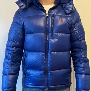 Blå dunjacka från Ralph Lauren - Säljer en snygg blå dunjacka från Polo Ralph Lauren. Jackan har en dragkedja framtill, långärmad design och en praktisk huva. Perfekt för kyliga dagar med sin quiltade stil och ikoniska logga på bröstet.