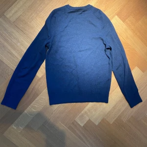 Blå merinoulltröja från Morris - Säljer en stilren blå tröja från Morris i 100% merinoull. Tröjan har en liten broderad logga på bröstet och långa ärmar. Perfekt för kyligare dagar.