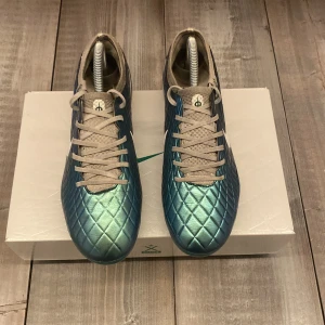 Blå fotbollsskor från Nike - Snygga blå fotbollsskor från Nike med quiltad yta och vit logga på sidan. Skorna har snörning och en slitstark sula med dobbar för bra grepp på planen. Perfekta för fotbollsspelare som vill ha stil och funktion. Priset är inte hugget i sten!!!