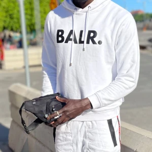 Vit hoodie från BALR. - Säljer en stilren vit hoodie från BALR. med svart logotyp på bröstet. Hoodien har en klassisk design med dragsko i huvan och långa ärmar. Perfekt för en avslappnad look.